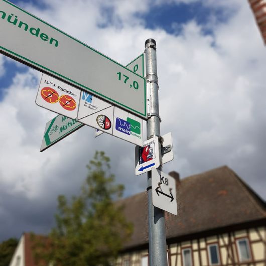 Auf einer Kreuzung neben einem Fachwerkhaus in Karlstadt steht ein Wegweiser. Er weist den Weg nach Würzburg, Mühlbach – und trägt das Schild des Marienwegs. 